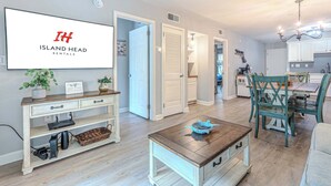 Un salon moderne avec un téléviseur diffusant des images de « Island Head Rentals », un coin repas avec des chaises de couleur turquoise et une cuisine dotée de meubles blancs.