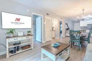 A modern living room with a television displaying 'Island Head Rentals,' a dining area with teal chairs, and a kitchen with white cabinets. - Beachfront Resort Villa Pool, Steps to the Sand (Hilton Head Island)