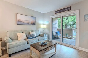 A living room with a sofa, coffee table, and a painting on the wall. There is a sliding glass door leading to an outdoor area with furniture. - Beachfront Resort Villa Pool, Steps to the Sand (Hilton Head Island)