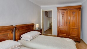 A bedroom with two beds, a wooden wardrobe, a nightstand with a lamp, and a door leading to another room.