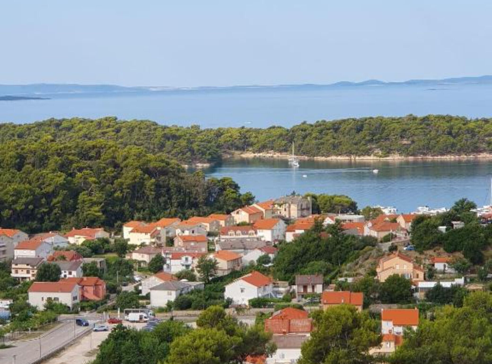 Une ville côtière aux bâtiments aux toits rouges, dotée d’un port de plaisance et bordée d’une colline boisée.