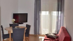 Quarto com televisão, mesa de jantar com cadeiras, sofá vermelho e janela com cortinas estampadas.