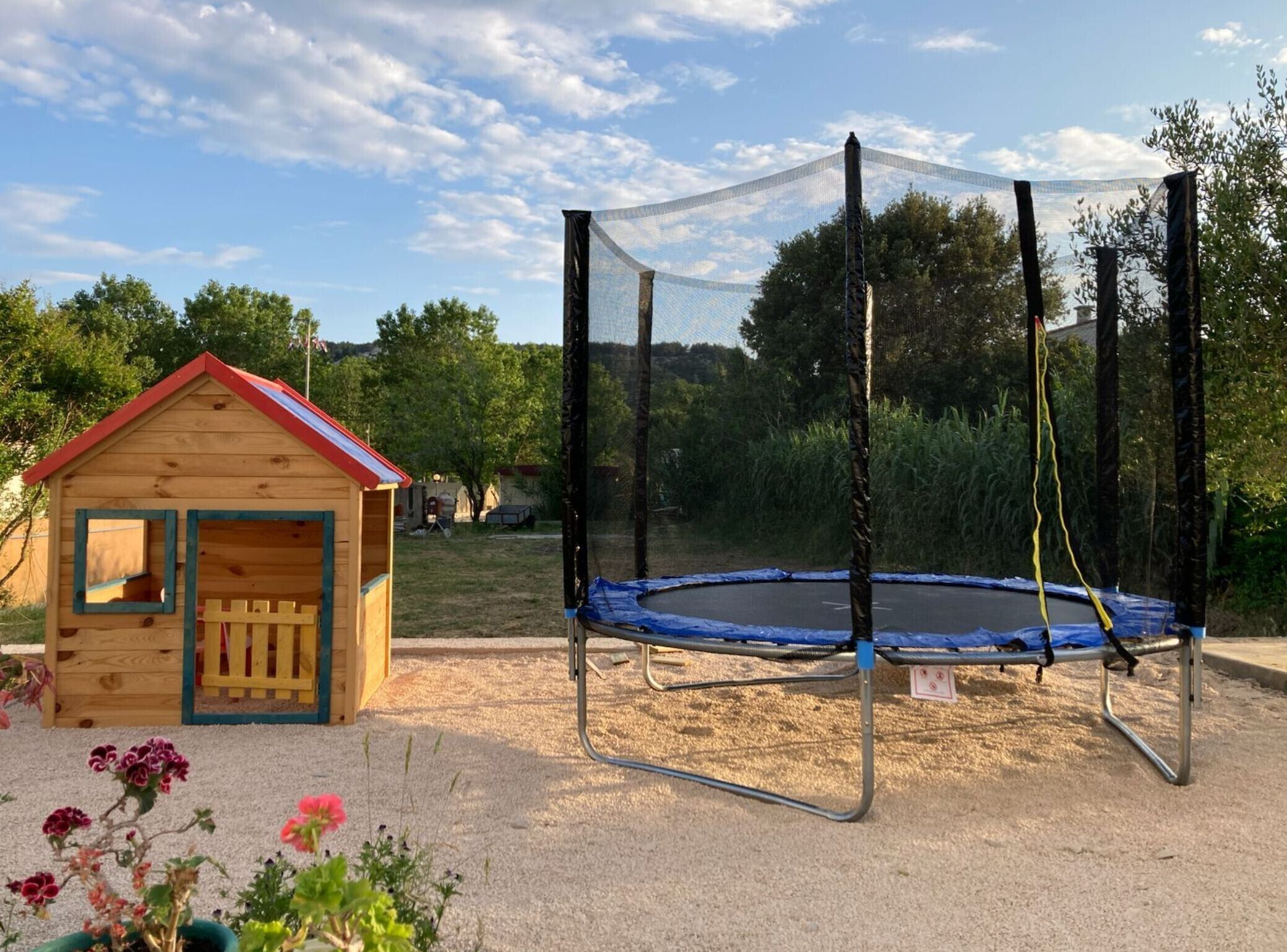 Une cabane en bois avec un toit rouge, un trampoline doté d’un filet de sécurité et un sol sablonneux.