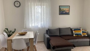 Un salon avec un canapé, une table à manger et des chaises, une horloge, une fenêtre avec des rideaux et un tableau accroché au mur.