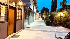 Une vue nocturne d’une rue bordée d’immeubles, avec un trottoir et quelques espaces verts.