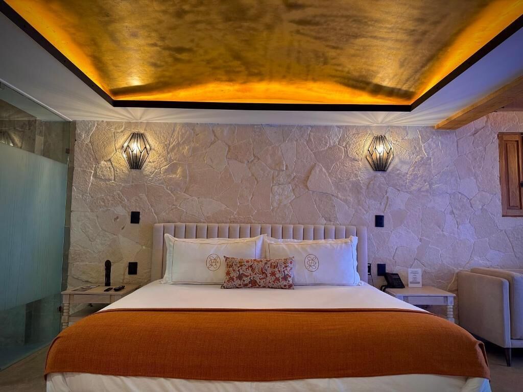 A bedroom with a stone wall, a bed with white bedding, two wall-mounted lights, and a wooden headboard.