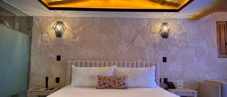 Un dormitorio con pared de piedra, cama con ropa de cama blanca, dos luces fijadas a la pared y cabecero de madera.