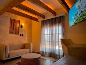A cozy living room with a sofa, a round ottoman, a wooden ceiling, a stone wall, a window with sheer curtains, and a painting. - Casa Laja - Hotel Boutique (San Miguel de Allende)