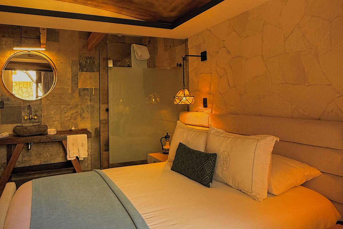 A hotel room with a bed, a round mirror, a nightstand, a lamp, and a stone wall.