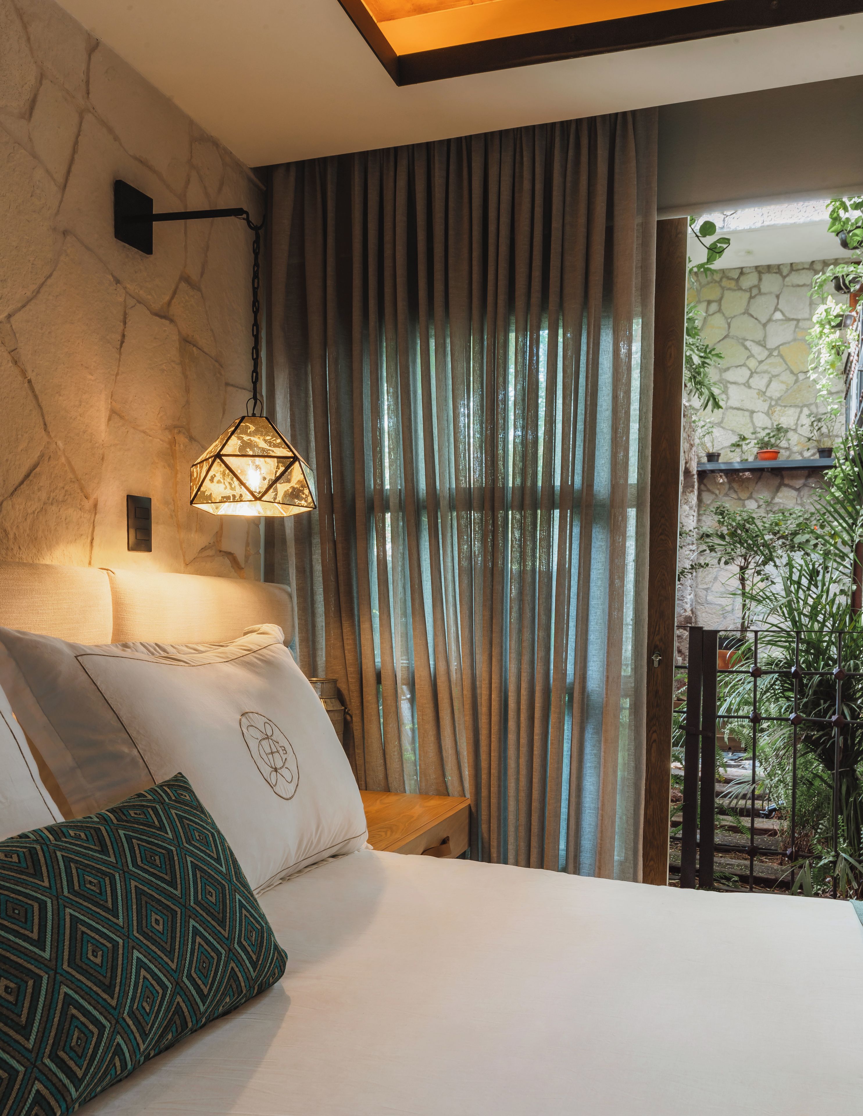 Ein Schlafzimmer mit Stehwand, einem Bett mit gemusterten Kissen, einer Hängeleuchte und Blick auf einen Garten durch ein Fenster.