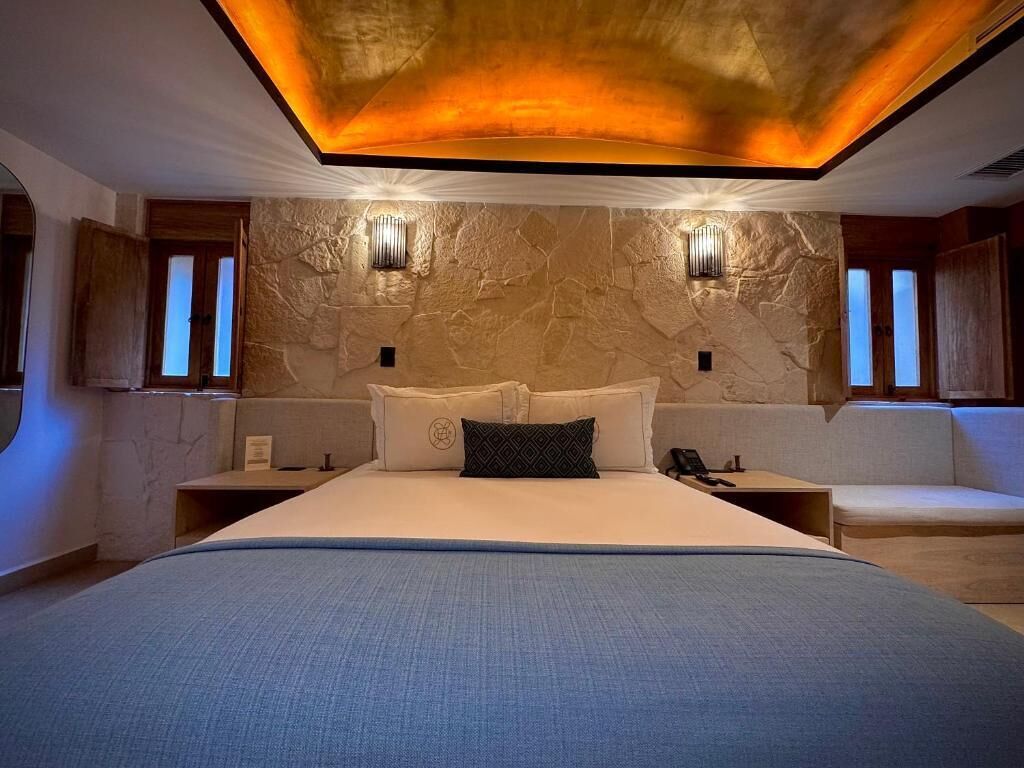 A modern bedroom with a large bed, two bedside tables, a headboard with built-in lighting, and a stone wall.