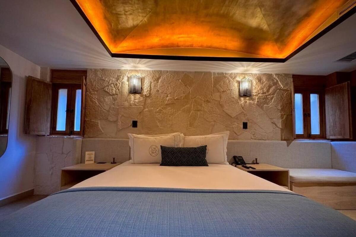 A modern bedroom with a large bed, two bedside tables, a headboard with built-in lighting, and a stone wall.