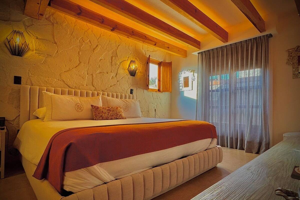 A bedroom with a large bed, stone wall, wooden beams, a window with curtains, and a nightstand.