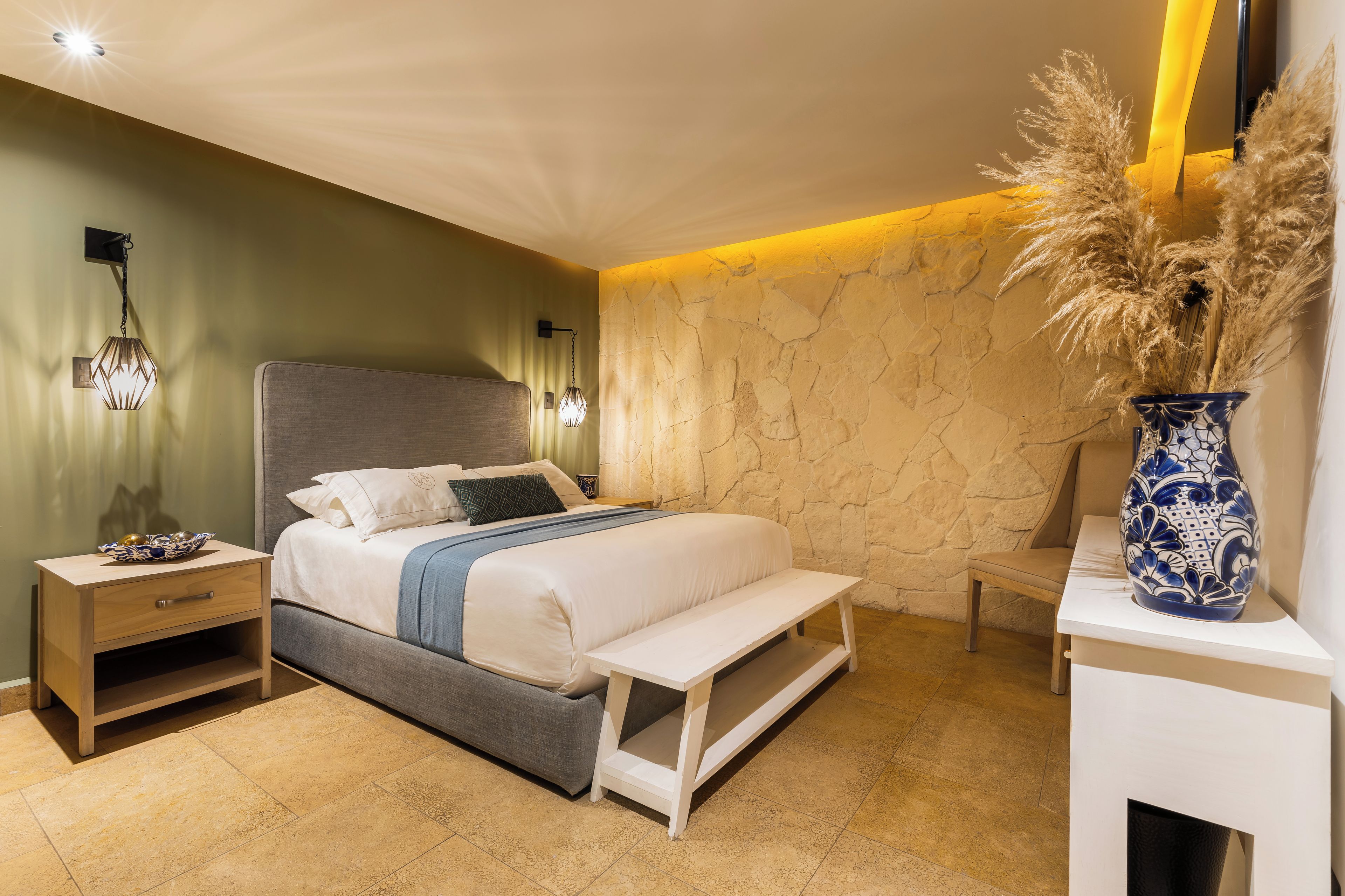 A modern bedroom with a bed, bedside tables, a nightstand, a bench, a vase with dried plants, and a textured wall.