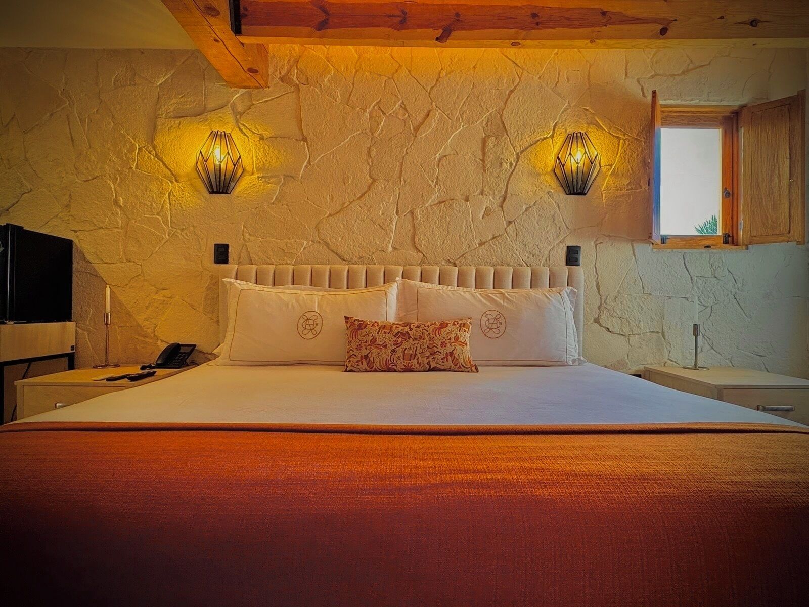 A bedroom with a stone wall, a bed with white bedding, two wall-mounted lamps, and a window with a view.
