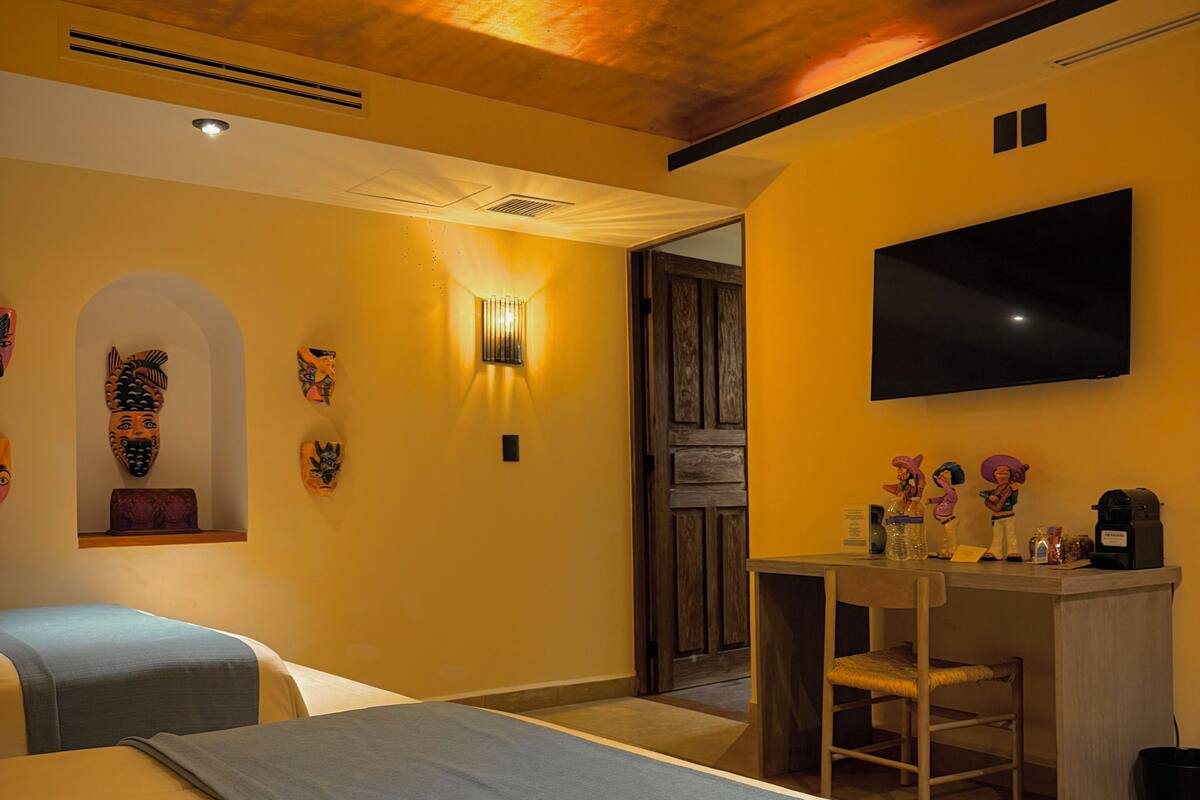 A hotel room with two beds, a desk, a chair, a TV, and decorative masks on the wall.