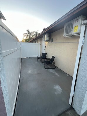 A covered outdoor patio with two lounge chairs, a white fence, and an air conditioning unit mounted on the wall.