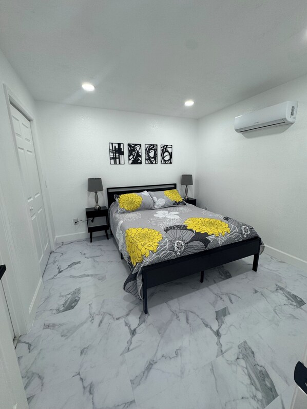 A bedroom with a bed, a nightstand, a lamp, a wall-mounted air conditioner, and a door.