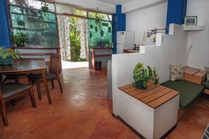 Een kamer met een eettafel, stoelen, een kleine bank, een koelkast en een raam met uitzicht op groen.