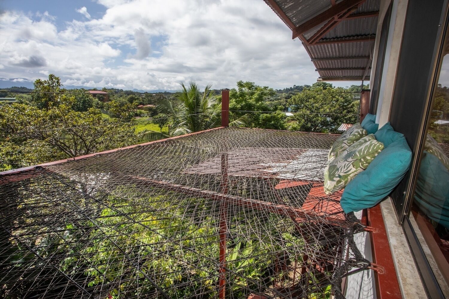 Ein Balkon mit Drahtgitter-Geländer, einem roten Dach und Blick auf eine grüne Landschaft mit Bäumen und Hügeln.