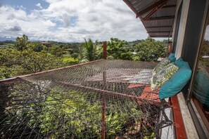 Een balkon met een hekwerk van gaas, een rood dak en uitzicht over een groen landschap met bomen en heuvels.