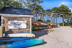 Ein Gebäude direkt am Strand, mit der Aufschrift 'Boat House' und einem Schild 'Point Check-In', umgeben von bunten Kajaks und einer Holzleiter.
