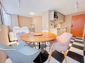 Ein Zimmer mit einem runden Holztisch, zwei Stühlen, einer Couch, einem Schachbrettfußboden, einer Küchenzeile und einer Tür.