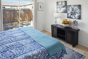 Ein Schlafzimmer mit gestreiftem Bettbezug, einem Nachttisch mit einer Vase voller Sonnenblumen und Blick auf eine Terrasse mit Stühlen und einem Zaun.