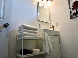 Ein Badezimmer mit einem weißen Regal, das gefaltete Handtücher beherbergt, einem Waschbecken mit Armatur, einem Spiegel und einer Wandleuchte.