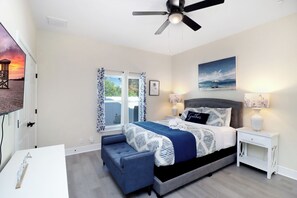 Quarto com cama, mesas de cabeceira, ventilador de teto, janela com cortinas e um quadro na parede.