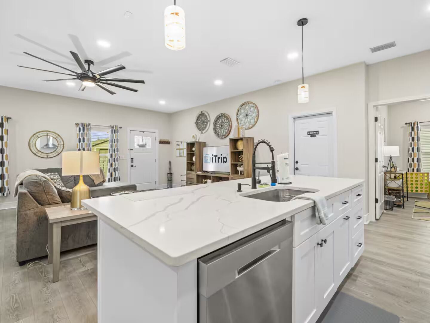 A modern kitchen with a central island, a ceiling fan, pendant lights, and a dining area with a sofa and lamp.
