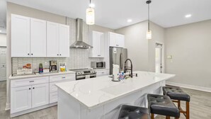 Una cocina moderna con armarios blancos, una isla grande con taburetes de bar, electrodomésticos de acero inoxidable e iluminación colgante.