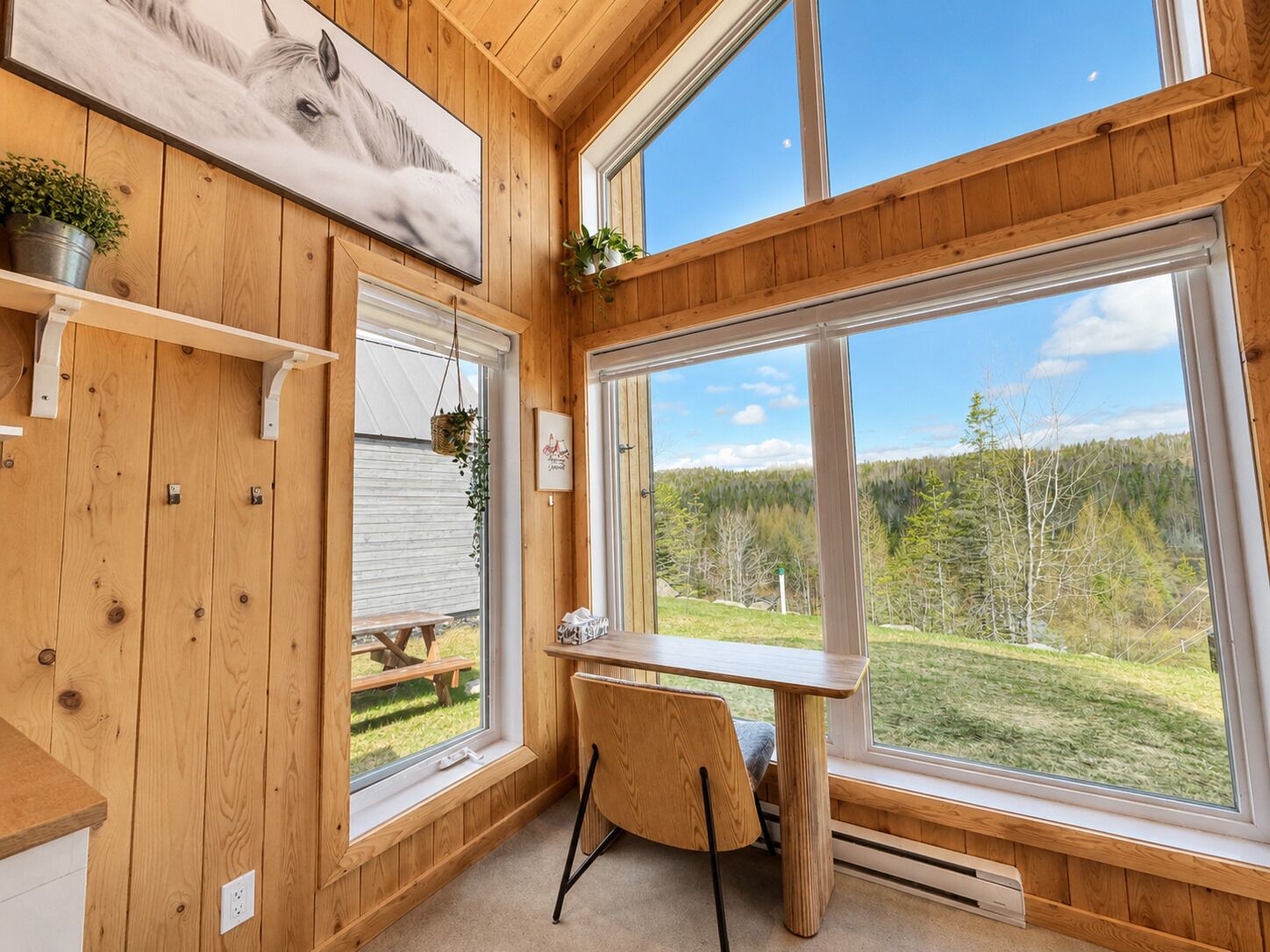 A cabin interior with wooden walls, a dining area, a large window with a view of a forest, and a mounted picture of a horse.