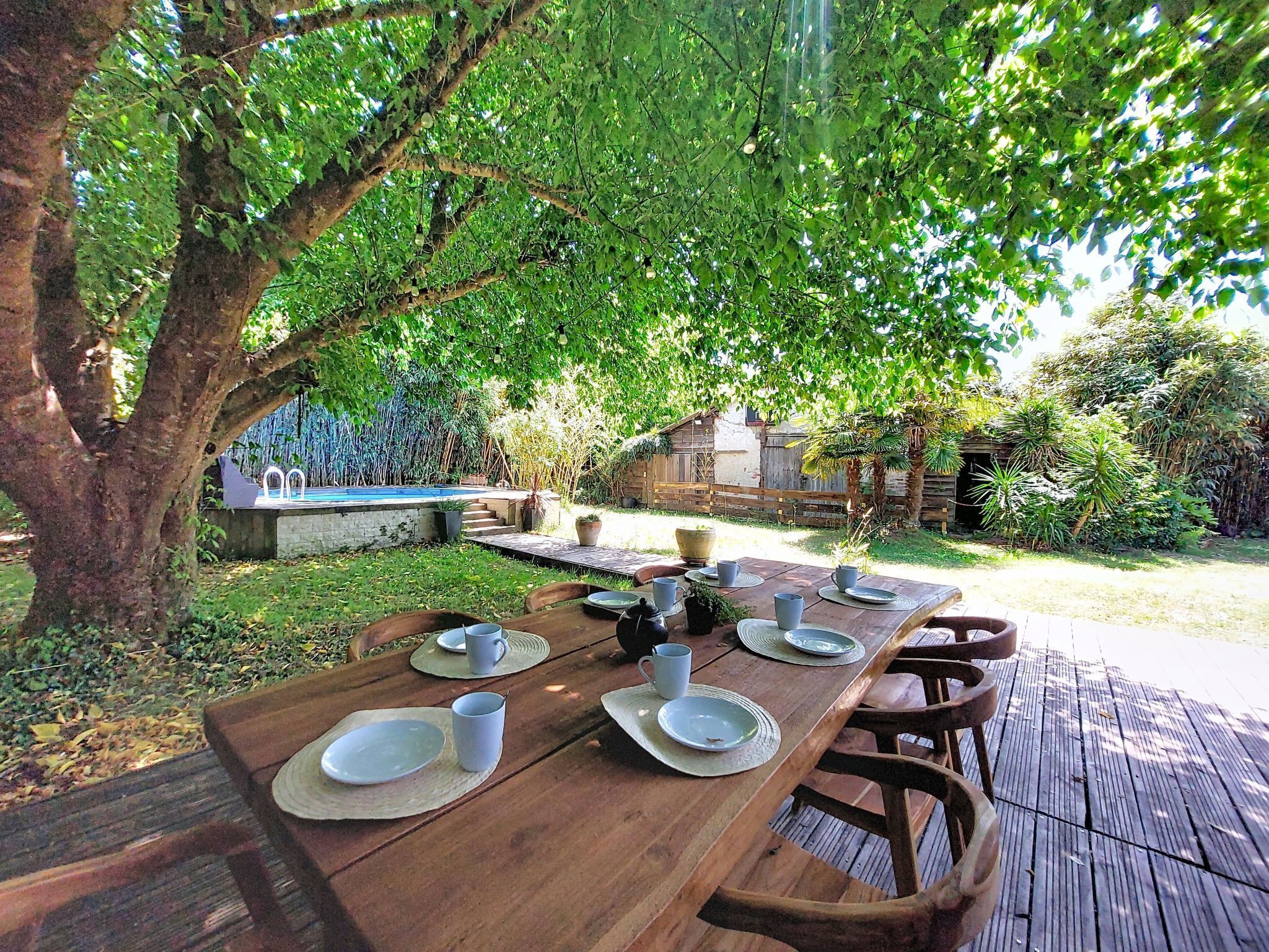 An outdoor dining area with a wooden table set for four, surrounded by chairs, under a large tree.
