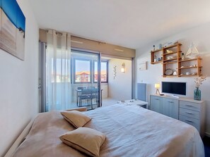 A bedroom with a bed, a desk, a TV, and a balcony with a view of buildings. - Le point d'Or by Interhome (Hossegor)