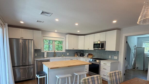A modern kitchen with stainless steel appliances, a wooden island, white cabinetry, and a dining area with a wooden table and plaid chairs.