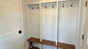 A room with a white door, wooden flooring, a black rug, and a white wall with shelves and hooks.
