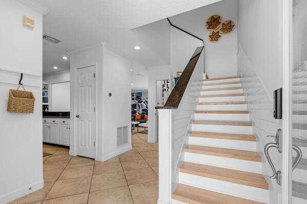 Una escalera de madera con barandilla, que conduce a una habitación con paredes blancas y una cocina visible al fondo.