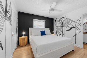 Un dormitorio con cama, ventilador de techo, mesita de noche con lámpara y ventana con persianas.