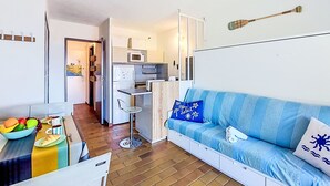 A compact living space with a kitchenette, a sofa with patterned cushions, a dining table with a bowl of fruit, and a small stool. - Les Aigues Marines by Interhome (Saint Cyr sur Mer La Madrague)