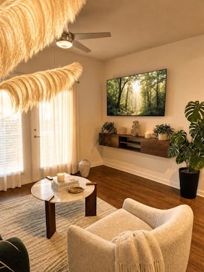 A living room with a sofa, a coffee table, a TV mounted on the wall, a ceiling fan, and a large window with curtains. - Modern Lakeview Oasis, King Bed • Gym • Pool (Jacksonville)