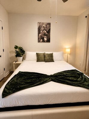 A neatly made bed with white linens and a dark green throw, a wall-mounted elephant picture, a bedside plant, and a lamp. - Modern Lakeview Oasis, King Bed • Gym • Pool (Jacksonville)