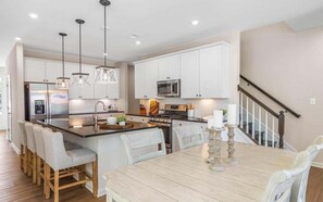 A modern kitchen with white cabinets, a central island, pendant lighting, and a dining area with a wooden table and chairs. - Bayside * 4BR Luxury Home w Incredible View (Selbyville)