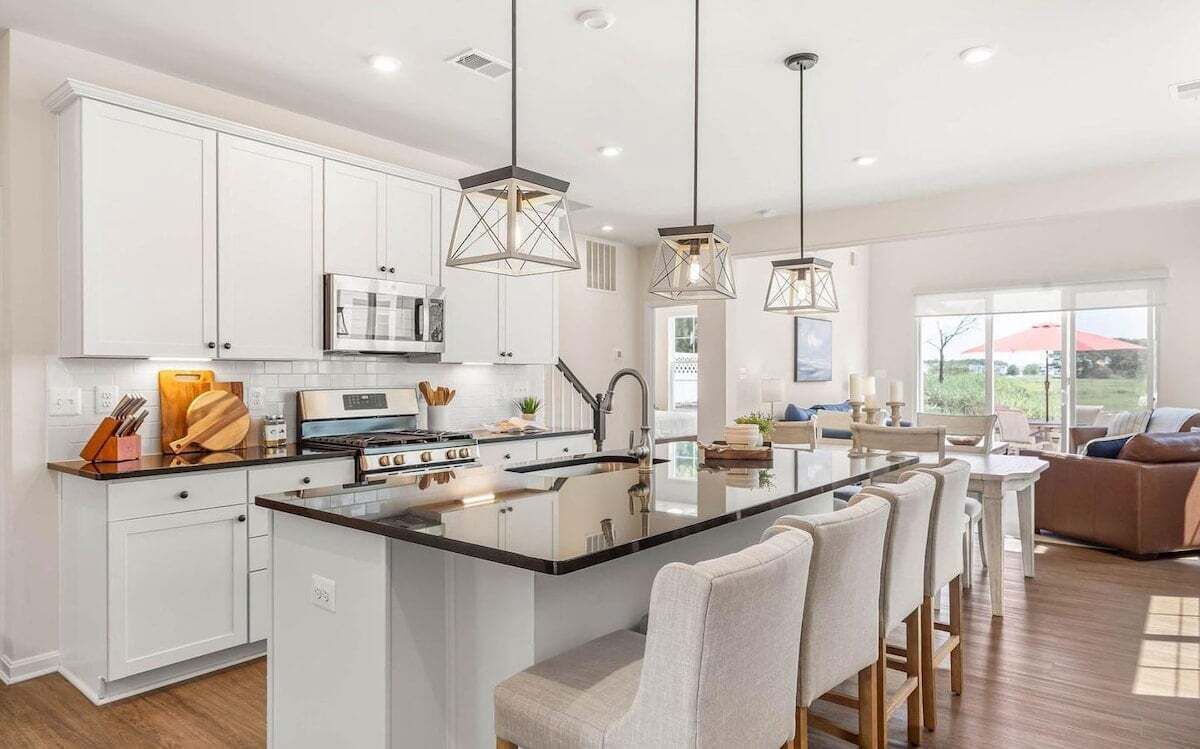 A modern kitchen with white cabinets, a central island, pendant lighting, and a dining area with chairs.