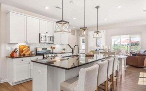 A modern kitchen with white cabinets, a central island, pendant lighting, and a dining area with chairs. - Bayside * 4BR Luxury Home w Incredible View (Selbyville)