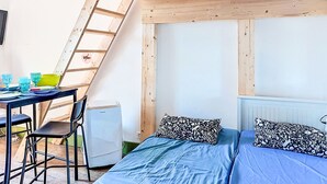 Quarto com teto estilo loft, cama com almofadas estampadas, mesa com cadeiras e radiador.
