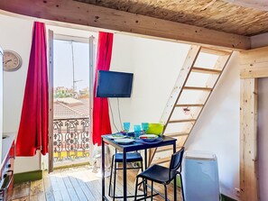 Ein Zimmer mit Holzboden, einem an der Wand befestigten Fernseher, einem Tisch mit Stühlen und einem Fenster mit roten Vorhängen.