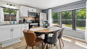 Une cuisine moderne avec des meubles blancs, un four à micro-ondes et un réfrigérateur. On y trouve une table à manger avec des chaises, et la vue donne sur un espace verdoyant.