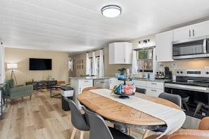 Una cocina moderna con armarios blancos, un microondas y una estufa. Hay un comedor con una mesa y sillas de madera.