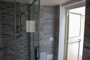 Une salle de bain moderne avec une douche à l’italienne en verre, des murs recouverts de carreaux gris et une porte blanche.